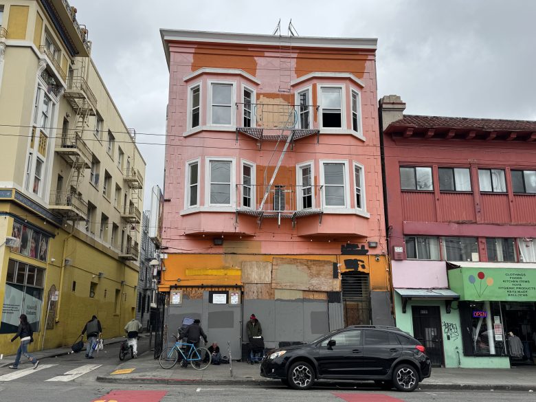 A three-story pink building with boarded-up windows on the ground floor stands between yellow and red buildings on a cloudy day; people and a black SUV are in front.
