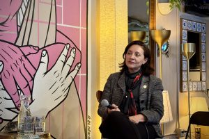 A woman sits on a chair holding a microphone, next to a table with glasses and a carafe, with a large illustrated mural and various lamps in the background.