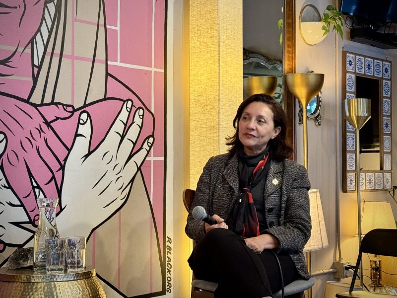 A woman sits on a chair holding a microphone, next to a table with glasses and a carafe, with a large illustrated mural and various lamps in the background.