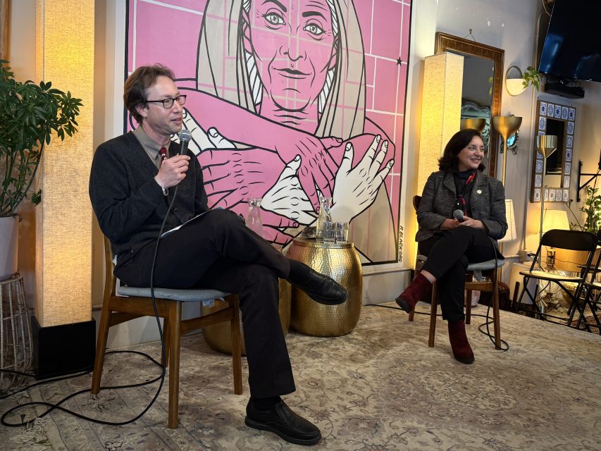 Two people sit on chairs and speak into microphones in a room with warm lighting and a large artwork of a woman with crossed arms behind them.