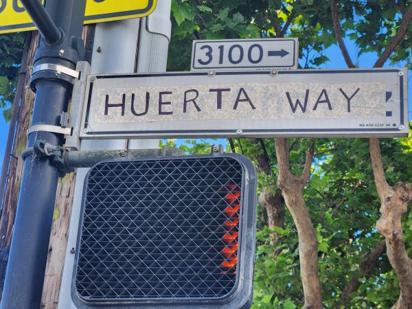 Street sign reading "HUERTA WAY" at the 3100 block, with a pedestrian crossing signal displaying a red hand. Trees and a pole are visible in the background.
