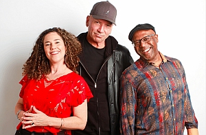 Three adults stand together smiling in front of a white background; one woman in red and two men, one wearing a cap and one with glasses and a patterned shirt.