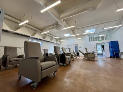 A room with several gray reclining chairs on wheels arranged in rows, fluorescent ceiling lights, and medical equipment visible in the background.