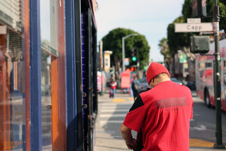 A person in a red shirt and hat stands on a city sidewalk near a street intersection with trees and buildings in the background.