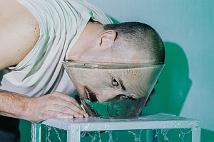 A person bends over a glass bowl filled with water, submerging their face, with water spilling over the edge.