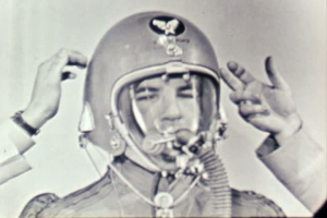 A person wearing a flight helmet and oxygen mask has their helmet adjusted by someone else, shown in a black-and-white photograph.