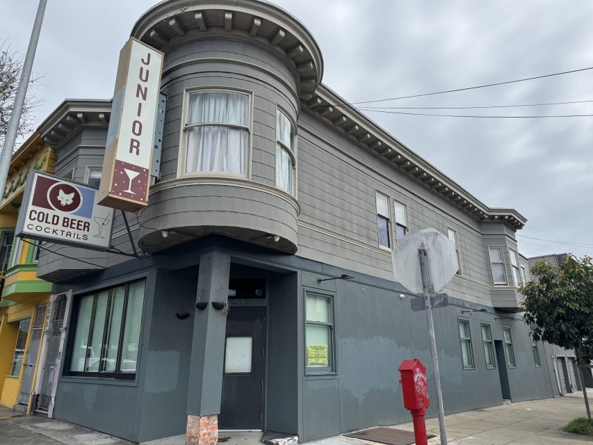 A gray, two-story corner building with a "Junior" sign and a "Cold Beer Cocktails" sign, located at a street intersection under a cloudy sky.