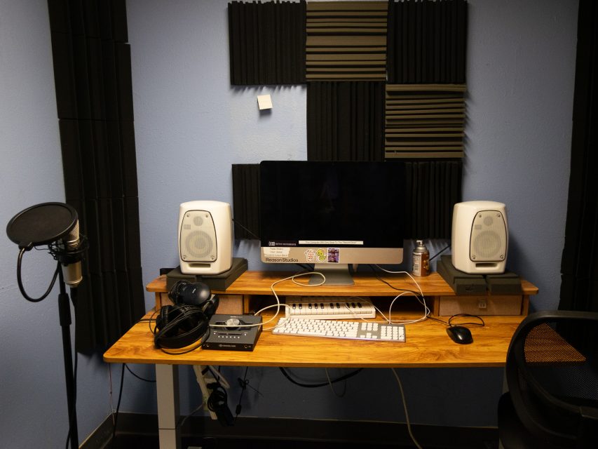 A recording studio setup with a computer, two speakers, a keyboard, microphone with pop filter, headphones, audio interface, and acoustic panels on a wooden desk.