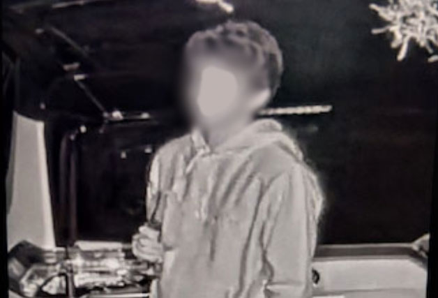 A person in a hoodie stands indoors holding an object, with their face blurred and background details dimly visible.