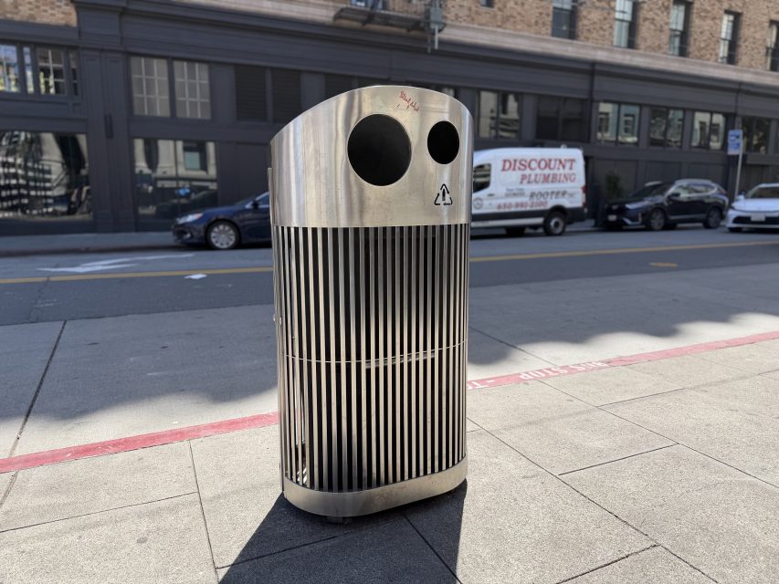 A modern stainless steel trash can stands on a city sidewalk near the curb; buildings and a white van are visible in the background.