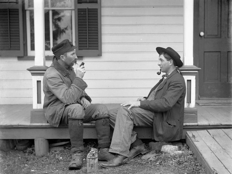 Two men sit on a porch; one drinks from a cup while the other smokes a pipe. A bottle sits on the ground between them. The house has wooden steps and a closed door.