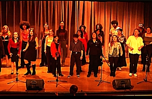 A group of people stands on a stage singing in front of microphones with a brown curtain backdrop.