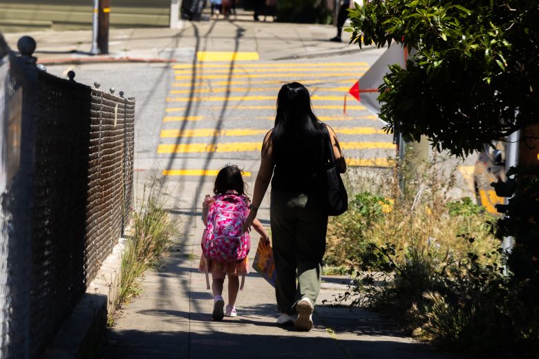 An adult and a child with a pink backpack walk hand-in-hand on a sidewalk toward a yellow-striped crosswalk on a sunny day.