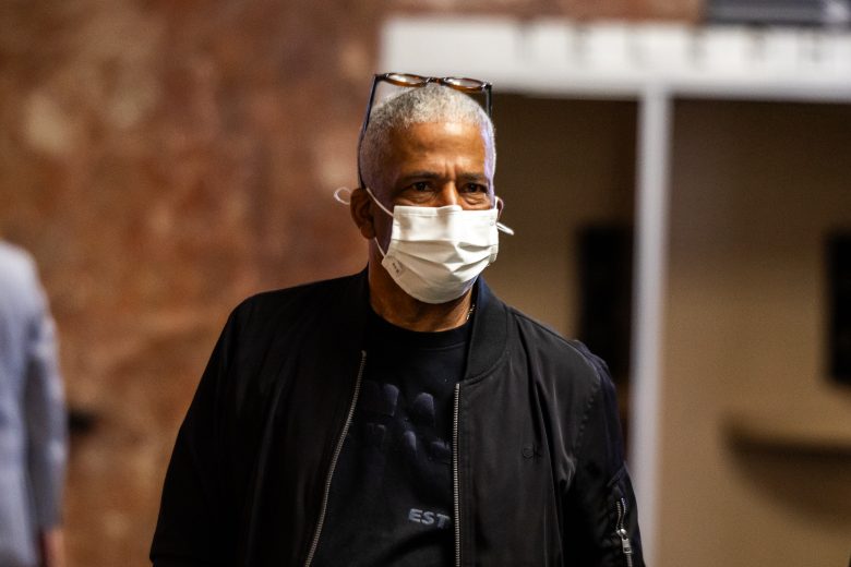 An older man with short gray hair wears a face mask, eyeglasses resting on his head, and a black jacket while standing indoors.