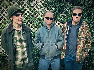 Three men stand outdoors in front of a white lattice fence and greenery, wearing casual jackets and sunglasses.
