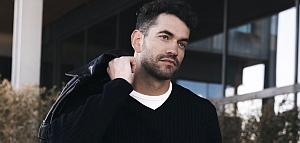 Man with short dark hair and beard wearing a black sweater and white shirt, holding a jacket over his shoulder, standing outdoors near a modern building.
