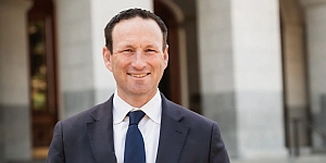 A man in a suit and tie stands outside a building with columns, smiling at the camera.