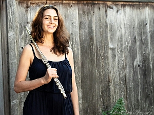A woman in a black sleeveless dress stands in front of a wooden fence, holding a flute and smiling at the camera.