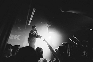 A performer on stage gestures towards an enthusiastic crowd with raised hands in a dimly lit venue.