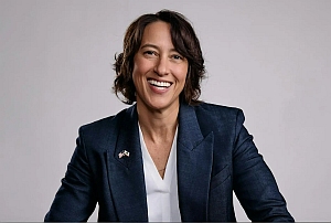 Person with short brown hair wearing a navy blazer and white shirt, smiling and seated against a plain light background.