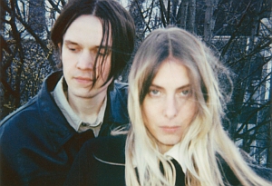 A man and a woman with long hair stand close together outdoors, with bare tree branches in the background.