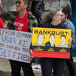 People holding protest signs about AI; one sign warns employees about being fired, another criticizes tech leaders with bold labels and claims tech is harmful for humanity.