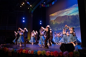 A group of dancers in blue costumes and hats perform a choreographed routine on stage with colorful lights, decorations, and a scenic backdrop.