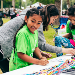 Three children sit at a table outdoors, making crafts with colorful strings at a community event.