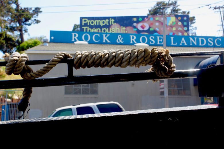 Coiled rope tied to a black metal bar in the foreground, with a sign reading "ROCK & ROSE LANDSC" and a colorful billboard above in the background.