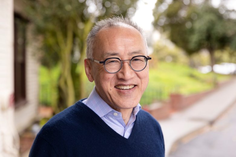 An older man with glasses and short gray hair smiles outdoors, wearing a blue sweater over a collared shirt. Trees and a sidewalk are visible in the blurred background.