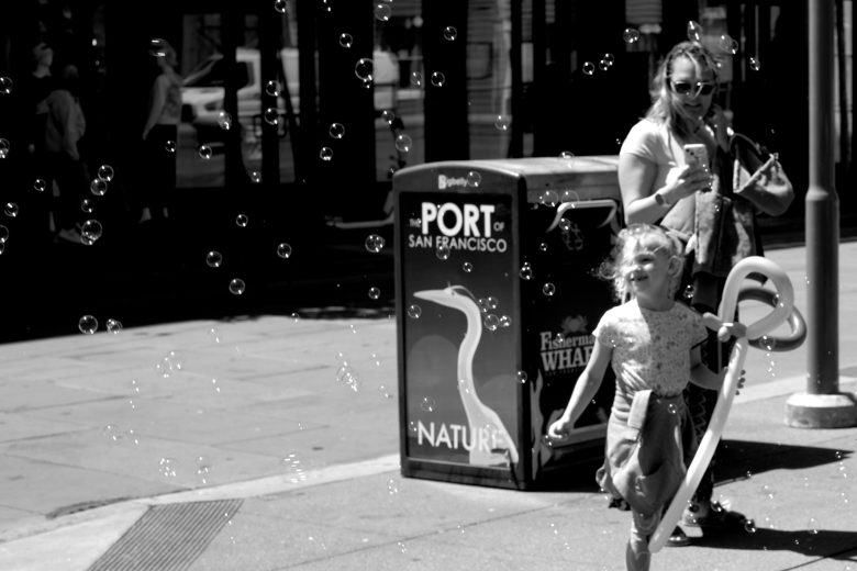 A young girl runs through bubbles near a woman holding a balloon animal on a city sidewalk by a "Port of San Francisco" trash bin.