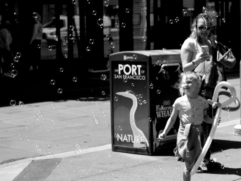 A young girl runs through bubbles near a woman holding a balloon animal on a city sidewalk by a "Port of San Francisco" trash bin.