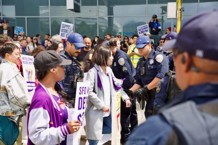 Police officers stand among a crowd of protesters holding signs during a demonstration outside a building labeled SFO.