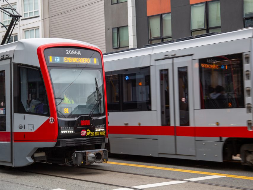 Two modern light rail trains pass each other on city tracks in front of contemporary apartment buildings.