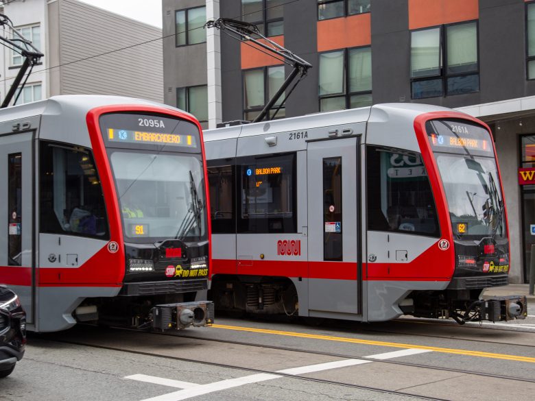 Two modern light rail trains stopped side by side on city tracks in front of mixed-use buildings.