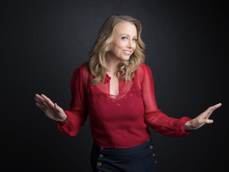 A woman with blonde hair in a red blouse and dark pants stands against a black background, smiling and holding her hands up as if gesturing.