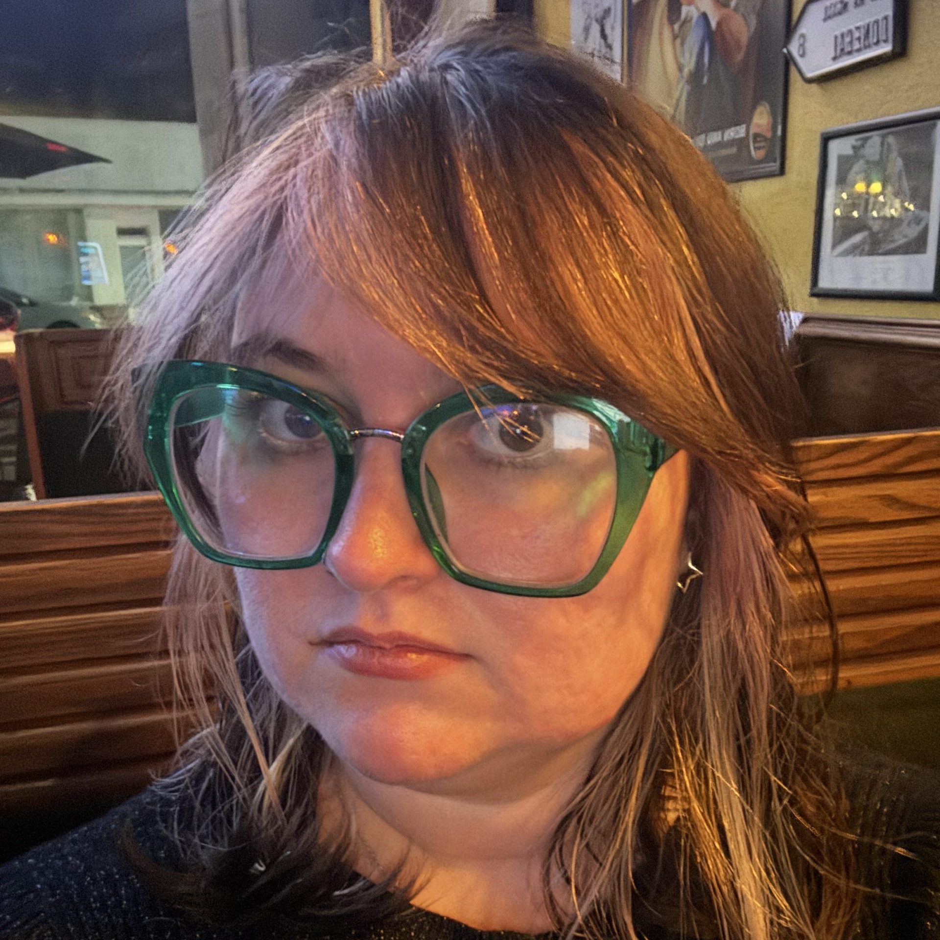 Person with long brown hair, wearing large green glasses and a black top, sits indoors with framed pictures on the yellow wall behind them.