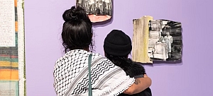 Two people stand closely together, viewing black-and-white photographs displayed on a light purple wall in an art gallery.