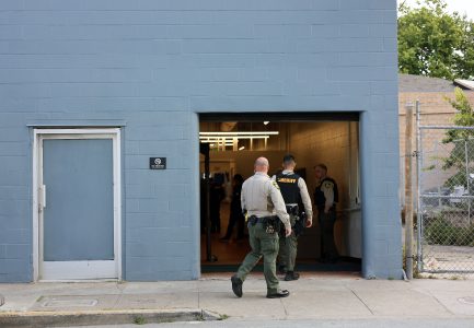 Two sheriff officers walk into a building with a blue cinderblock exterior, while other officers stand inside near an open garage-style door.