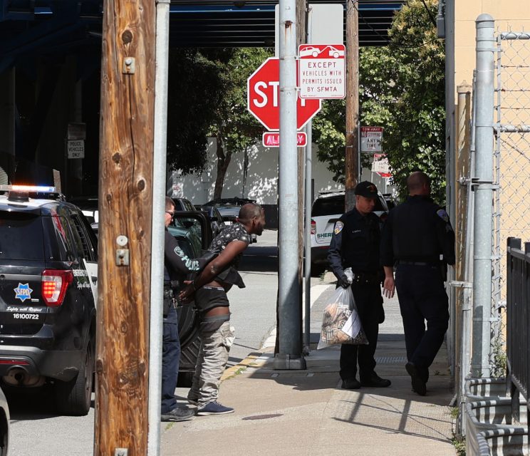 Two police officers stand near a police vehicle while a handcuffed man is being detained on a sidewalk by another officer, with a stop sign visible in the background.