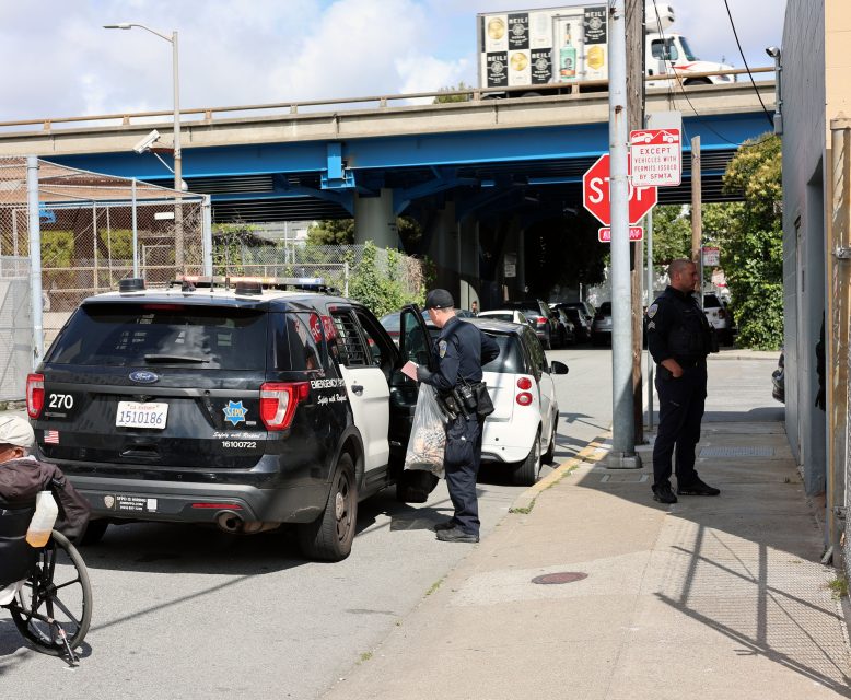 Two police officers stand beside a police SUV with its door open near a stop sign on a city street, while another person in a wheelchair passes by.