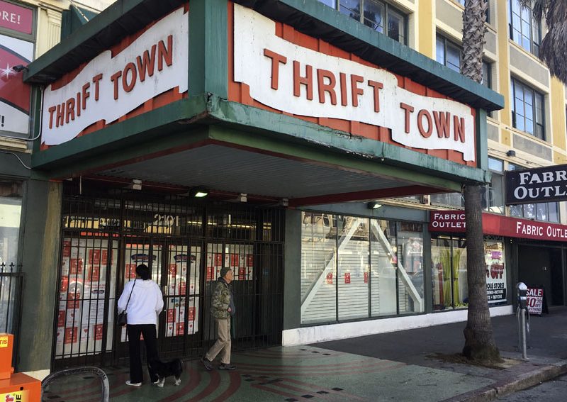 Two people stand outside Thrift Town, a thrift store with red and white signage promising 17 reasons to browse, next to a Fabric Outlet on a city street.