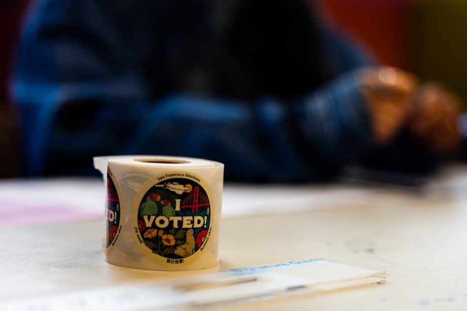 A roll of "I Voted!" stickers sits on a table, with a person's blurred hands and a pen visible in the background.