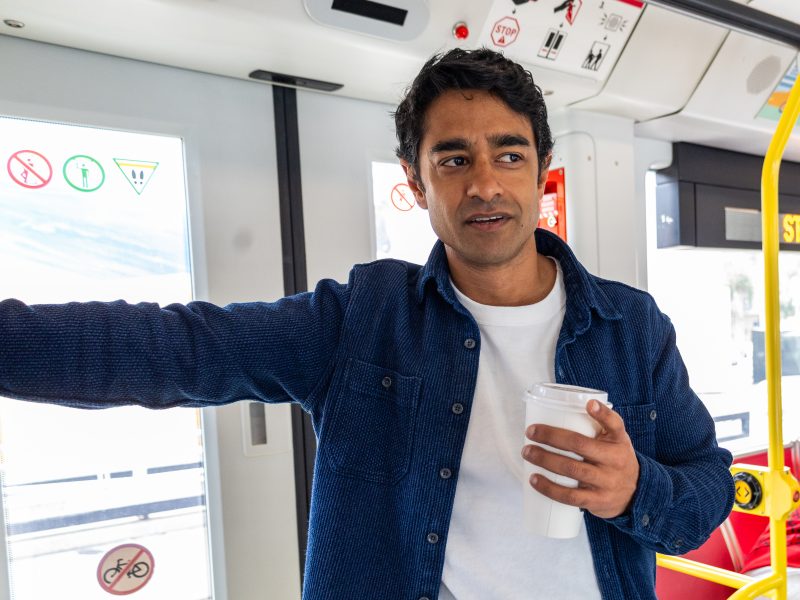 A man stands inside a bus holding a coffee cup in one hand and supporting himself with the other arm, next to doors marked with various no-entry symbols.