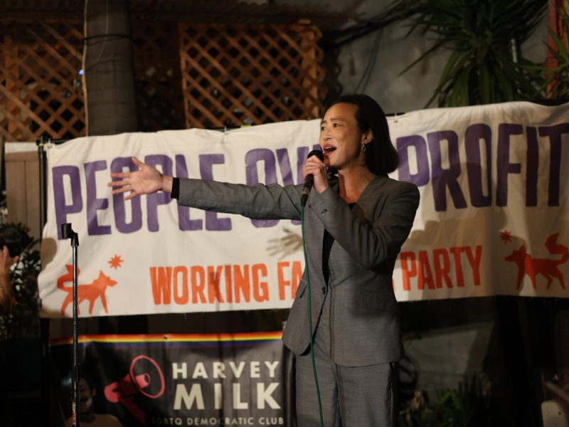 A person in a suit speaks into a microphone on stage in front of a sign that reads "People Over Profit" and "Harvey Milk LGBTQ Democratic Club.