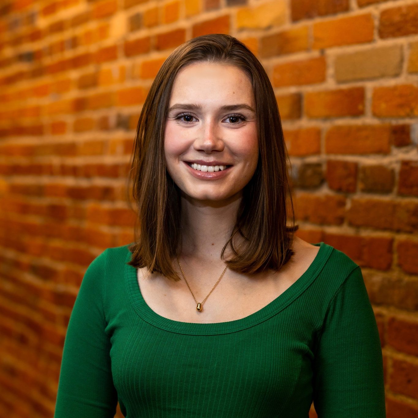 A woman with straight brown hair wearing a green top and a necklace smiles, standing in front of a brick wall.