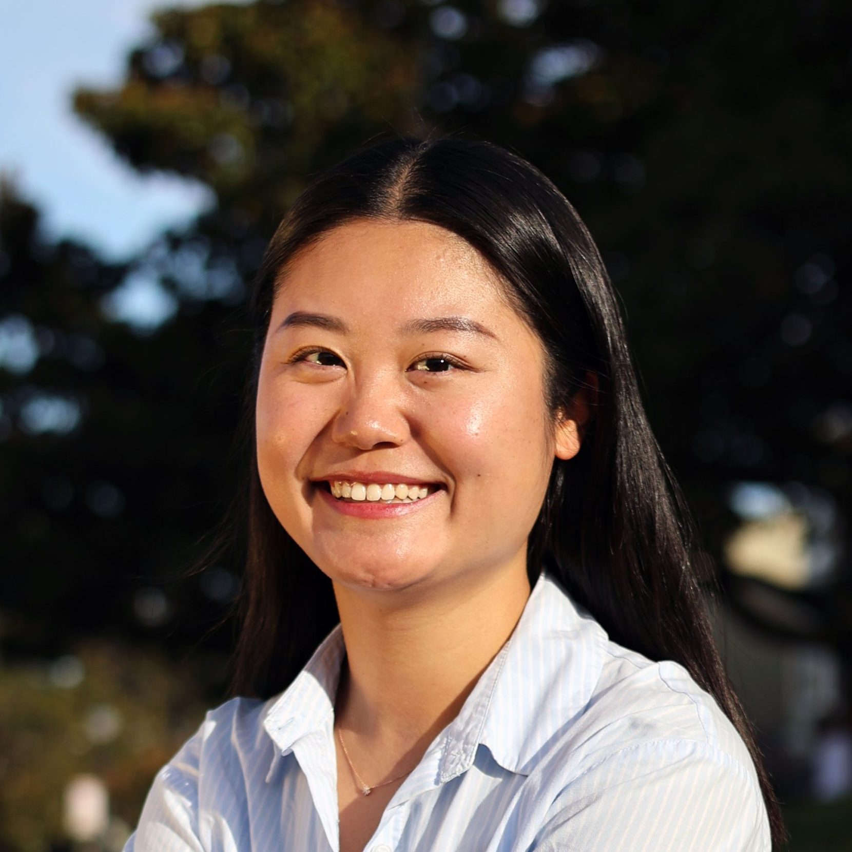A young woman with long black hair smiles outdoors, wearing a light blue collared shirt. Trees and a bright sky are visible in the background.