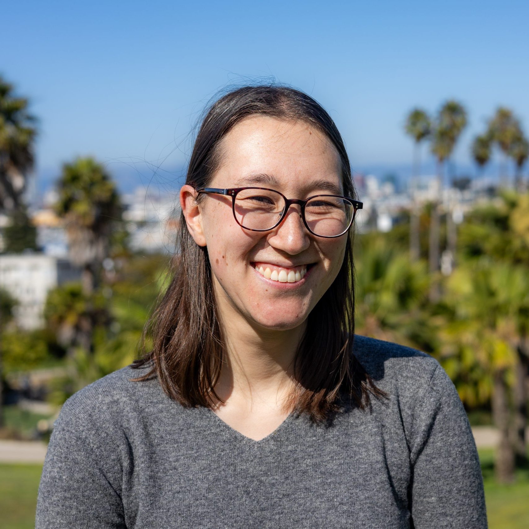 Person with glasses and long brown hair, wearing a gray sweater, smiling outdoors in a park with palm trees and a cityscape in the background.