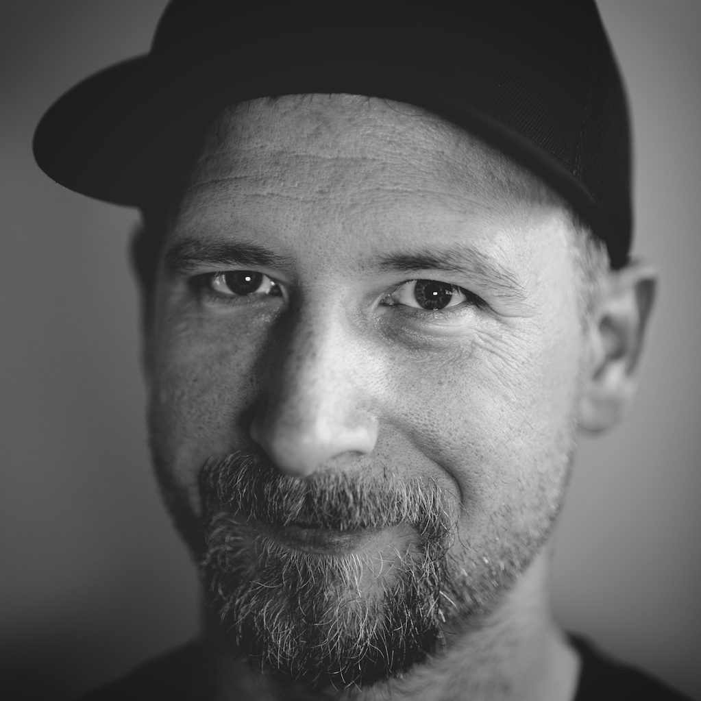Black and white close-up portrait of a bearded man wearing a dark cap, looking directly at the camera with a neutral expression.