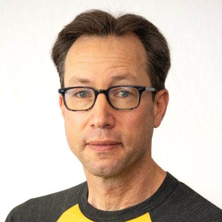 A man with short brown hair and glasses, wearing a black and yellow shirt, faces the camera against a plain white background.